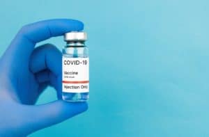 Mão com luva hospitalar de cor azul segurando frasco de vacina do covid