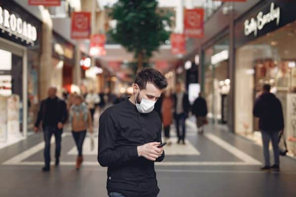 Rapaz usando máscara parado no meio de um local movimentado e olhando para o celular